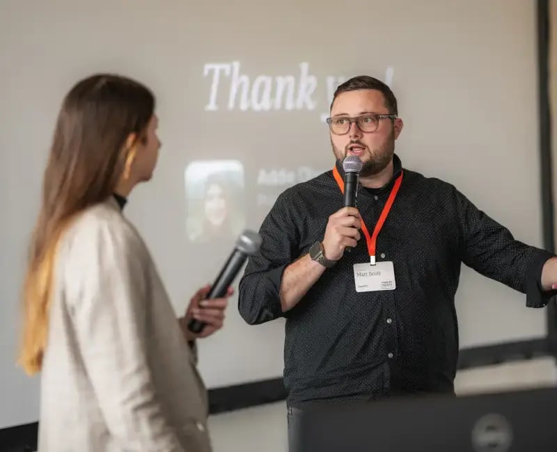 Two speakers hold microphones and speak in front of a presentation deck that says 