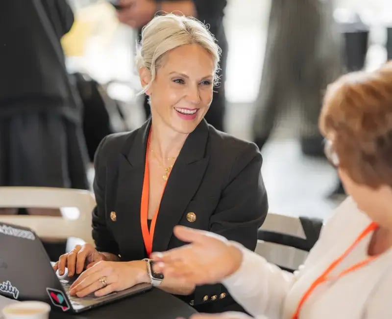 Two women sit at a table smiling. One woman uses a laptop.