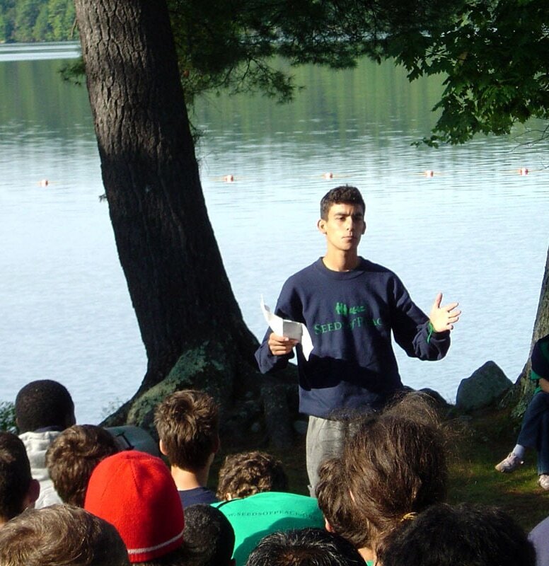 A man speaks to a group of youth by a lakeside